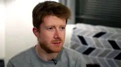 A man with short styled ginger hair and facial hair is looking to his left. He is wearing a brown round neck jumper and has a clip mic on the collar