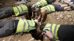 Kevin Church/BBC News Five archaeologists lying face down on the ground clustered around a grave. They are wearing bright yellow high-visibility vests and reaching down into the grave with their hands and tools.