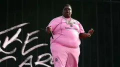 The rapper wearing a pink outfit with 'balenciaga' written on the chest, and several necklaces, holds a microphone in his extended right hand away against a black background with some chalk-like letters to his right 