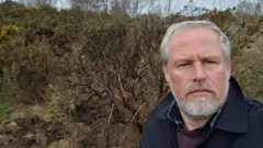 Durham County Councillor Mark Wilkes. He has short white hair and a trimmed beard. He has blue eyes and is wearing a blue coat, over a purple jumper. He is looking at the camera with a serious expression. Behind him are some gorse bushes.