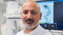 A man with short white beard wearing a white shirt stands in a medical room