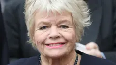 Judith Chalmers  smiling in a navy blue blazer and necklace in 2016
