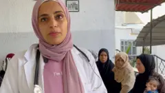 Dr. Sukar, pink handkerchief, pink T-shirt, white doctor's jacket and a stethoscope on the neck, looks into the camera when she answers questions in front of some patients