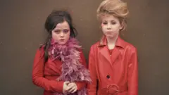 Nia Day as a younger child and Poppy. They are two white girls both wearing all red clothes and coats. Nia, left, has a reddish feather boa around her neck. Both girls have beehive type hairstyles and are made up.