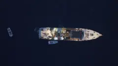 James Glancy/Martin Stalker The image is captured by a drone and is looking directly down upon a research vessel - with crew and equipment visible on deck - and two smaller dinghies close by. The picture is taken at dusk and the sea surrounding the vessel looks dark, inky blue