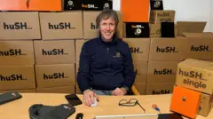 Hush Paul Nugent is sitting at a wooden desk behind which are stacks of cardboard boxes with Hush printed on them.