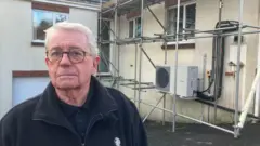 A man with glasses is looking straight at the camera - he has short white hair and is wearing a black fleece. Behind him is house with scaffolding surronding a heat pump which is mounted on the wall.
