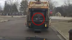 Man is hiding in a trash can with a garbage truck behind him.