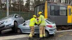 A training exercise that makes it look like a grey and yellow metro train has hit two grey cars at a level crossing. Two firefighters, who are wearing bright yellow helmets, are working on the nearest car.