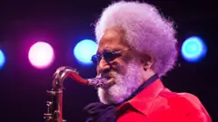 Getty Images Sonny Rollins plays saxophone while wearing a red jacket on stage under blue and pink lights.