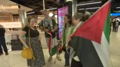 Four people are standing in an airport waiting area. One woman has a black tshirt and yellow bag, a man is holding a Palestine flag in his hands, two other women are smiling at the first girl. The woman to the extreme right is holding a large Palestine flag on a pole.