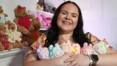Meg, a young woman with long dark hair grins as the camera while holding six colourful Labubu dolls in different pastel colours. There are teddy bears behind her and a shelf of other colourful soft toys, which are slightly blurred in focus.