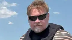 Karl Bushby A man with brown hair and grey beard is wearing a wet suit and sunglasses. Behind him is a blue sky.