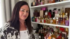 A woman with long dark brown hair wearing a cow print jacket standing in front of a display of perfumes