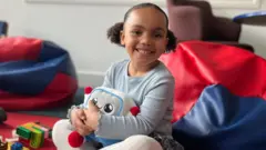 Picture of a young girl hugging Gabbo an AI-powered toy. She is sat, smiling among soft seating and other toys.