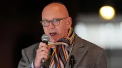 PA media Lord Wolfson speaking into the microphone. He is a bald man wearing glasses and a gray coat with a striped scarf.