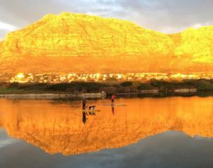 Two people take their dogs out for a paddle in a marina as the golden sun lights the mountain in the background