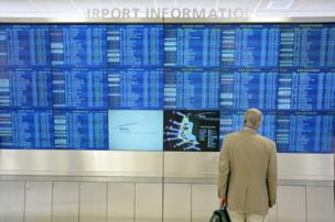 A person stands in front of a collection of airport information screens displaying arrivals and departures