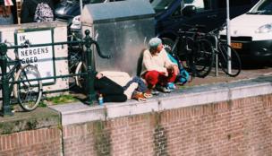 A woman is lying on the floor covering her face as she sleeps while a man squats next to her, smoking a cigarette