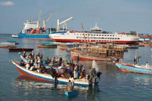 An large shipping container is contrasted with smaller boats carrying goods to supply the local market