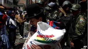 A woman carries a bag of sugar bought at a government subsidized market in La Paz as riot police stand guard