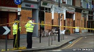 Police stand guard in front of boarded up shops and businesses in Ealing