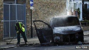 A street cleaner sweeps up around a smouldering van set alight during riots in Hackney in London