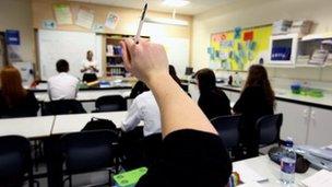 Pupils raise their hands in a classroom as a teacher stands by a whiteboard