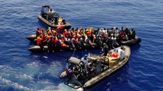 Small boats are pictured attending to migrants on top of an inflatable boat they are loaded on to.