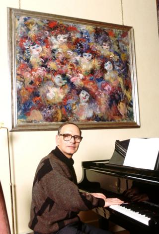Ennio Morricone sitting at a piano with a large painting on the wall