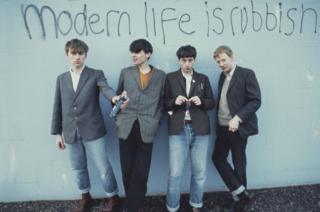 positive news Blur's four members holding spray cans in front of graffiti on a wall reading 
