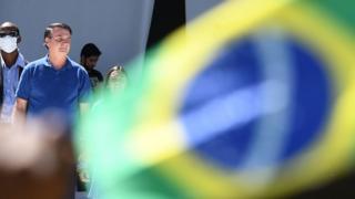 Bolsonaro appears behind the Brazilian flag and next to security with a mask