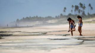 Duas banhistas na areia manchada de óleo, uma delas olhando para a sola do chinelo