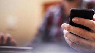 شخص يمسك بهاتفه المحمول بيد، ويضع الأخرى على لوحة مفاتيح جهاز الكمبيوتر