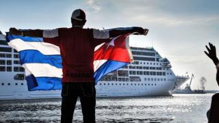 A man holds a Cuban flag with a cruise ship in the background