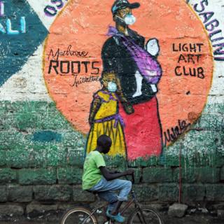 Ein Junge auf einem Fahrrad fährt vorbei an einem Wandbild einer Frau und eines Kindes in einer Gesichtsmaske im Slum Mathare, Nairobi, Kenia - Mittwoch, 8. April 2020