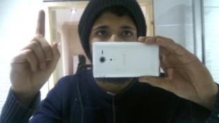 Aqib Imran taking a selfie and holding up one finger, in the style of an ISIS supporter