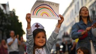 A girl dressed as a nurse holds up a drawing of a rainbow with the words "thank you" written on it