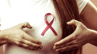 Mujer joven seálando a uno de sus senos donde lleva el lazo representativo de la lucha contra el cáncer de mama.