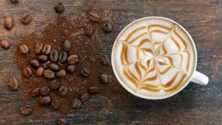 A cappuccino with a caramel web motif pictured next to a pile of coffee beans and grounds.