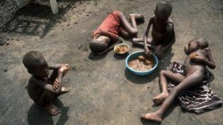 Four starving Biafran children sit and lay around a bowl of food in the dirt during the Biafran war