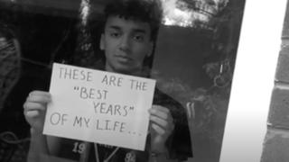 A young person holds up a sign that reads "these are the best years of my life"
