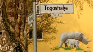 A street sign indicating a street named after the country of Togo is seen in front of a mural of a nursing rhinoceros on April 14, 2018 in Berlin, Germany.