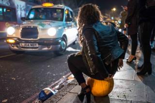 A woman sits on a pumpkin on a pavement next to a taxi