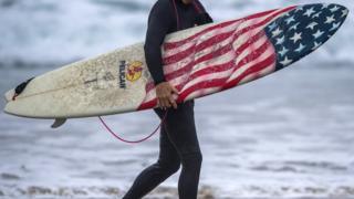 Серфер выносит свою патриотическую доску из воды после серфинга к югу от пирса Ньюпорт-Бич в Ньюпорт-Бич рано утром в пятницу, 7 сентября 2018 года,