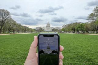 A mobile phone showing the time at noon in front of the United States Capitol, Washington, US