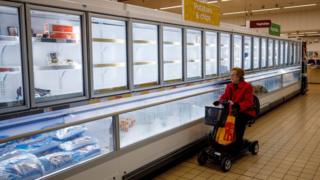 People shop in aisles with empty shelves in a Sainsbury