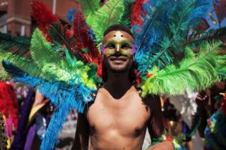 A man wearing a mask and a large collar of rainbow-coloured feathers walks alongside other people in the LGBTQ parade.