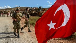 Турецкие солдаты стоят возле бронетехники, когда мужчина машет национальным флагом Турции во время демонстрации в поддержку операции «Идлиб» турецкой армии у границы между Турцией и Сирией возле Рейханлы, Хатай, 10 октября 2017 года