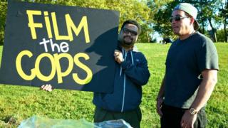Dennis Flores, um homem latino baixo de óculos, segura um cartaz escrito 'filme a polícia', em inglês, no meio de um parque